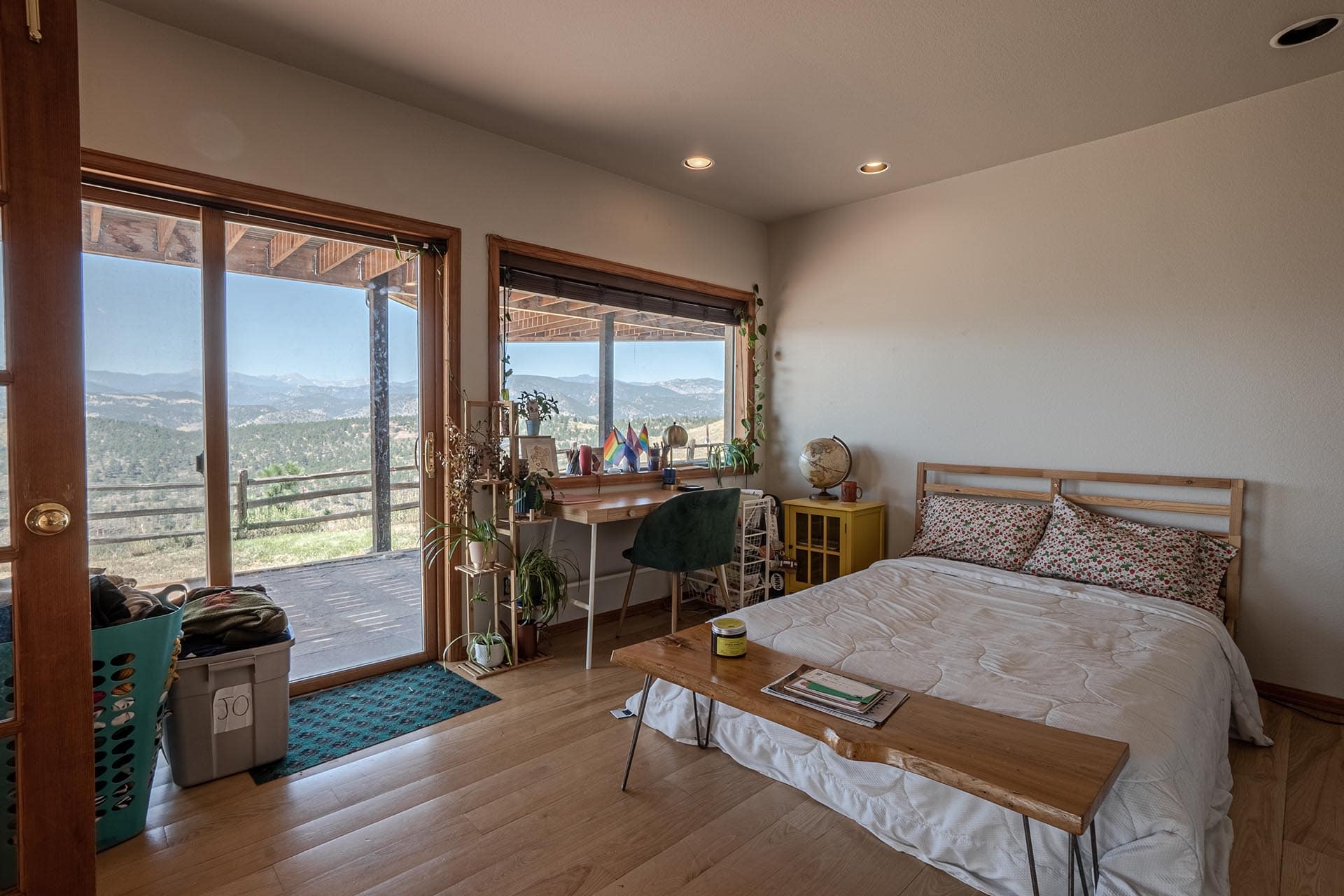 Bedroom with a bed and wooden bench, facing large windows that show a mountain landscape outside and a balcony door on the left.