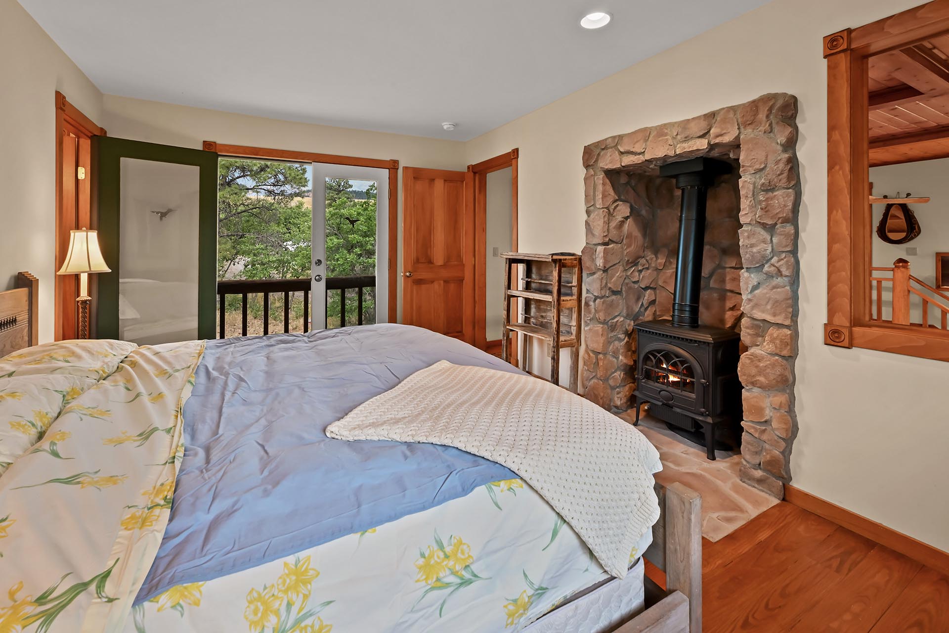 Cozy bedroom with a stone fireplace and wood stove, a bed with floral bedding, and a balcony door overlooking green trees outside.