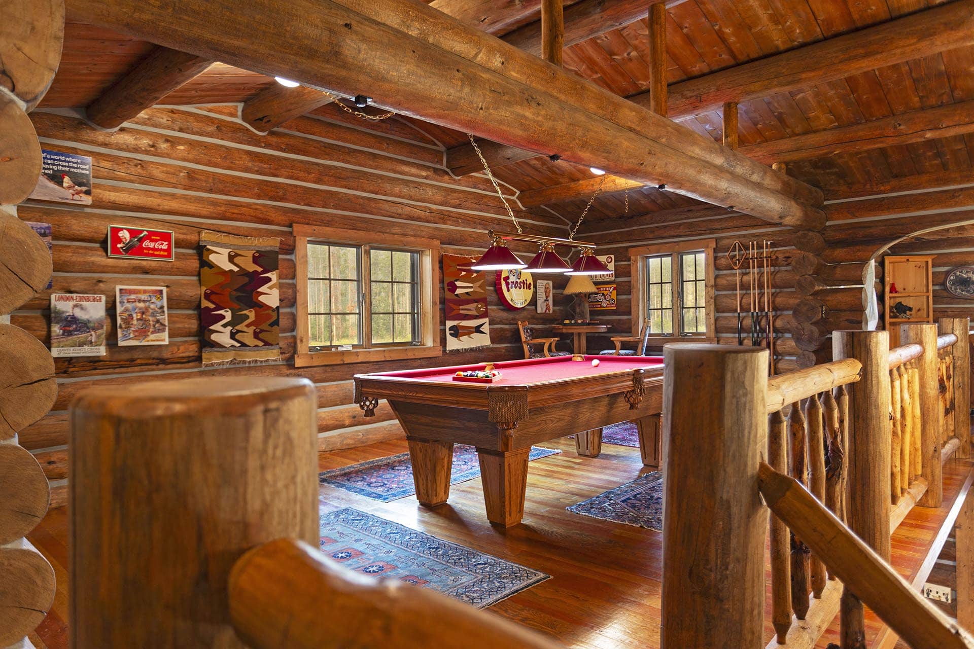 Rustic log cabin game room with a red billiards table under hanging lights and wooden beams.