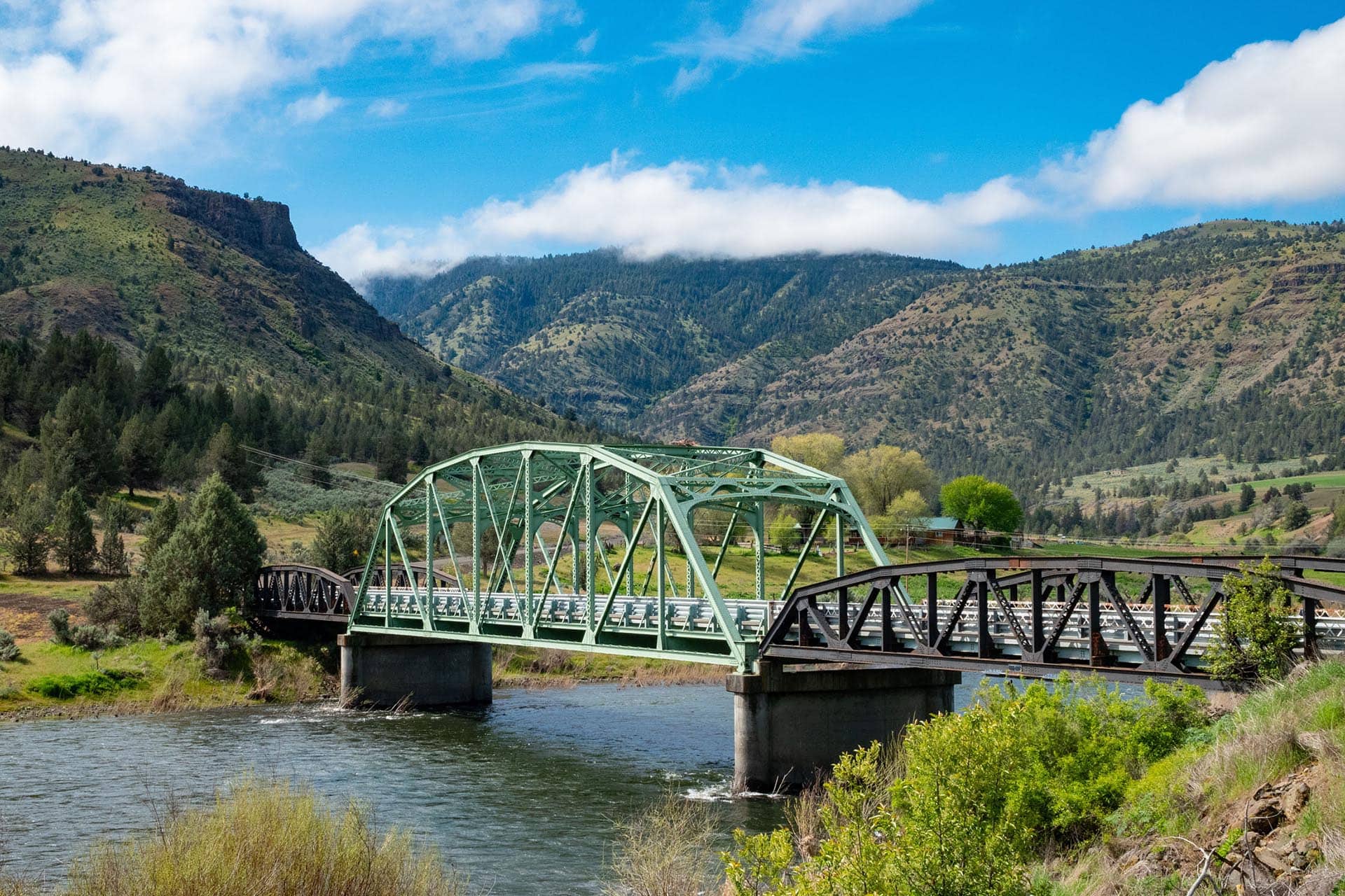Green steel truss bridge spans a calm river with forested hills and mountains under a bright blue sky with clouds in the background
