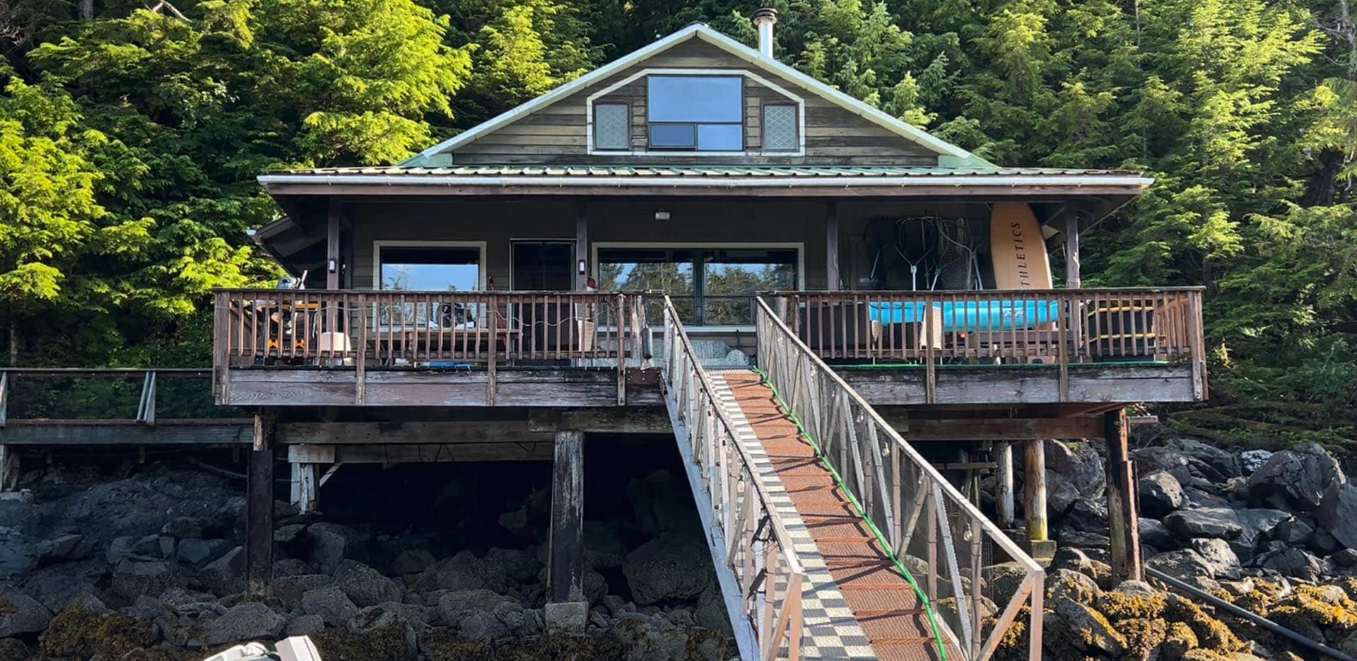 Wooden lakeside cabin on stilts with a long ramp leading to a deck over a rocky shoreline, backed by trees.