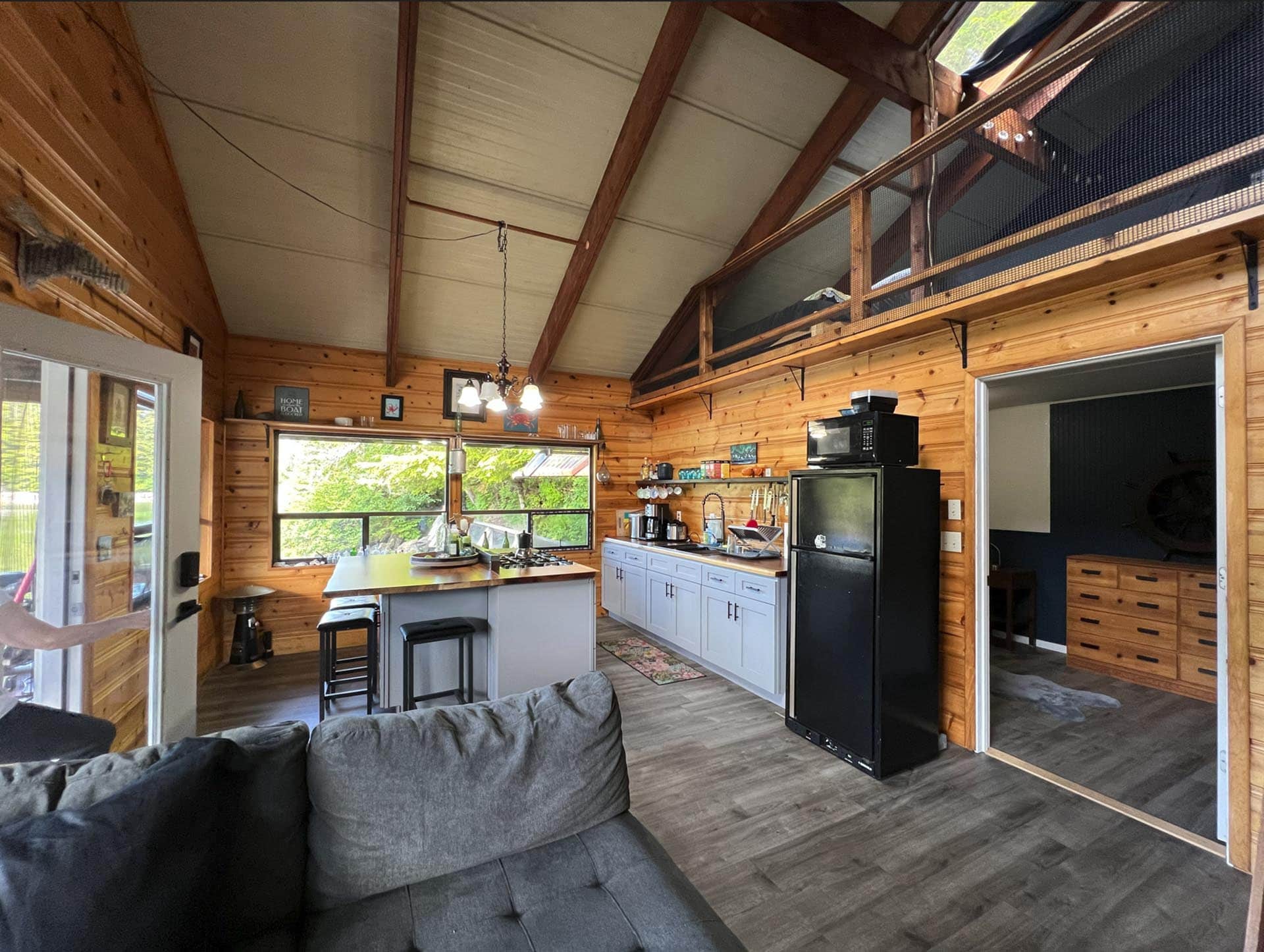Open-plan cabin kitchen and living area with a vaulted wooden ceiling, loft railing, and gray sectional sofa.