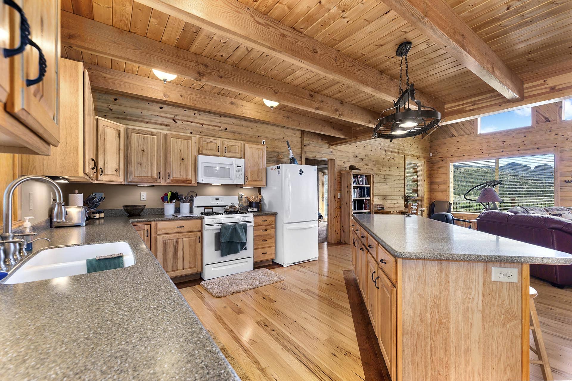 Rustic kitchen with wooden cabinets and a large island, stainless appliances, and exposed log beams in a cabin setting.