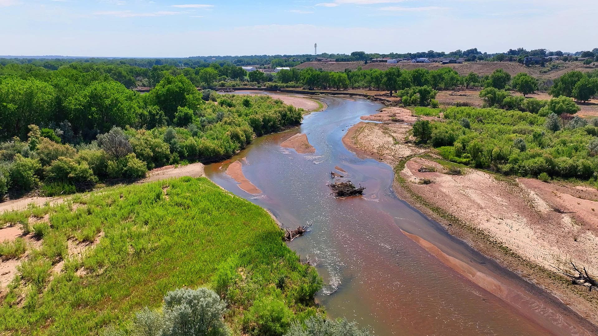 Aerial view of a winding river cutting through lush green trees and exposed sandy banks, with a distant rural town on the horizon.