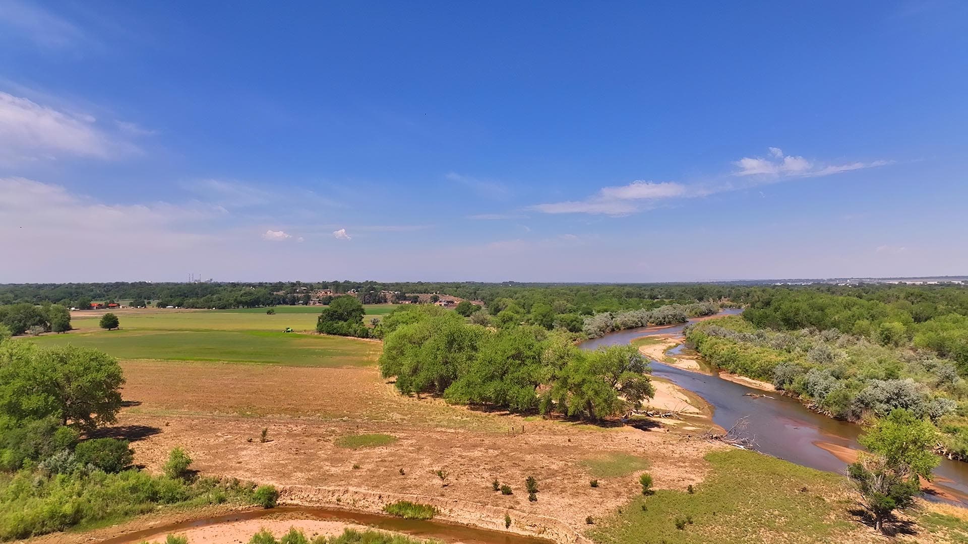 Wide rural landscape with a winding river, green trees, and fields under a clear blue sky.