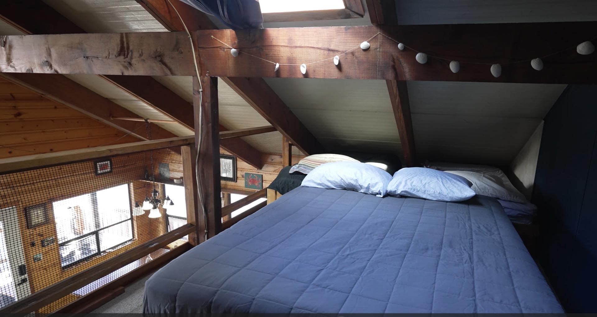 Loft bedroom with exposed wooden beams, string lights, and a blue quilted bed in foreground; railing and windows visible on the left.
