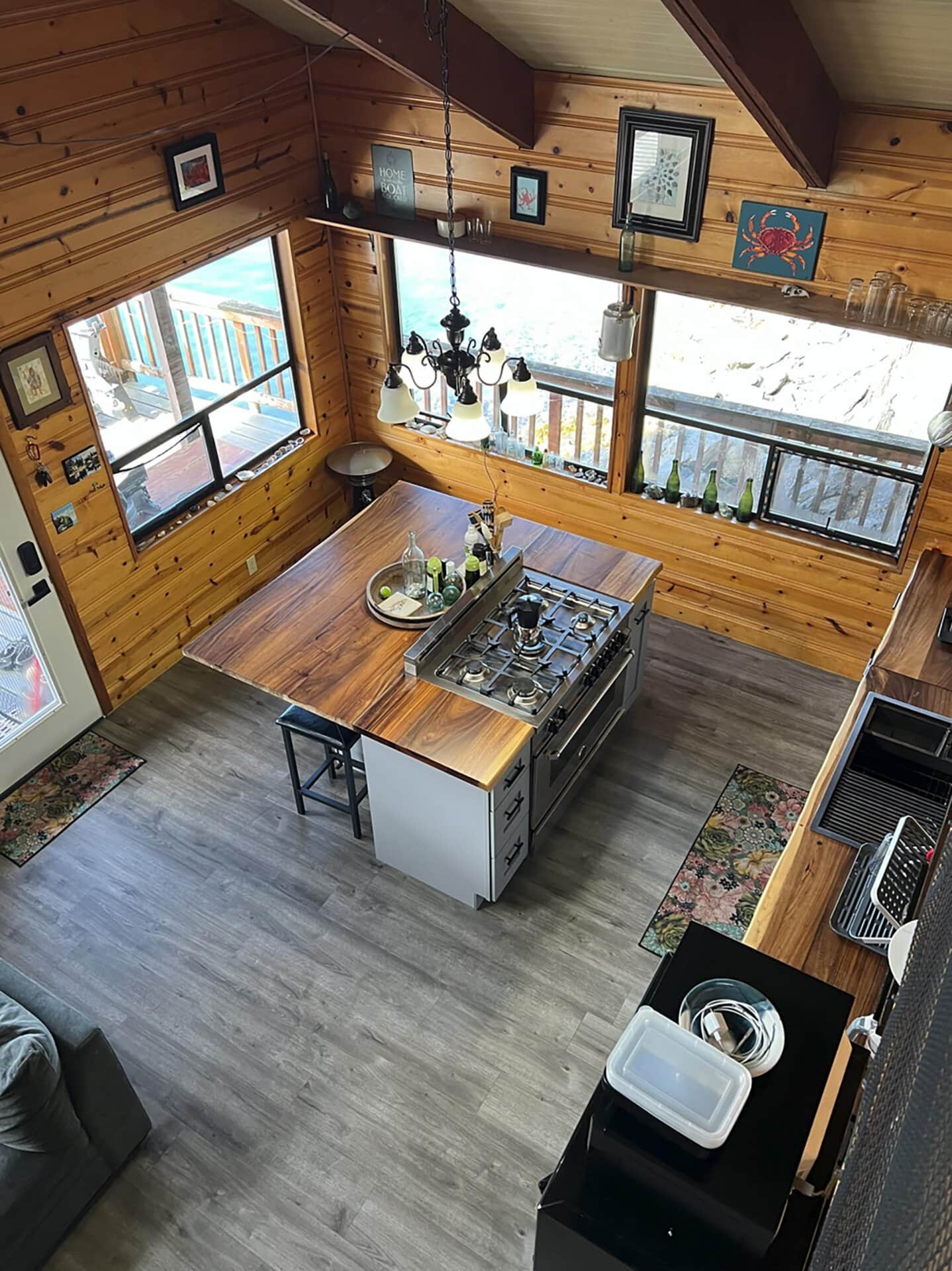 Wood-paneled kitchen with a central island gas stove, hanging chandelier, and large windows overlooking a deck.