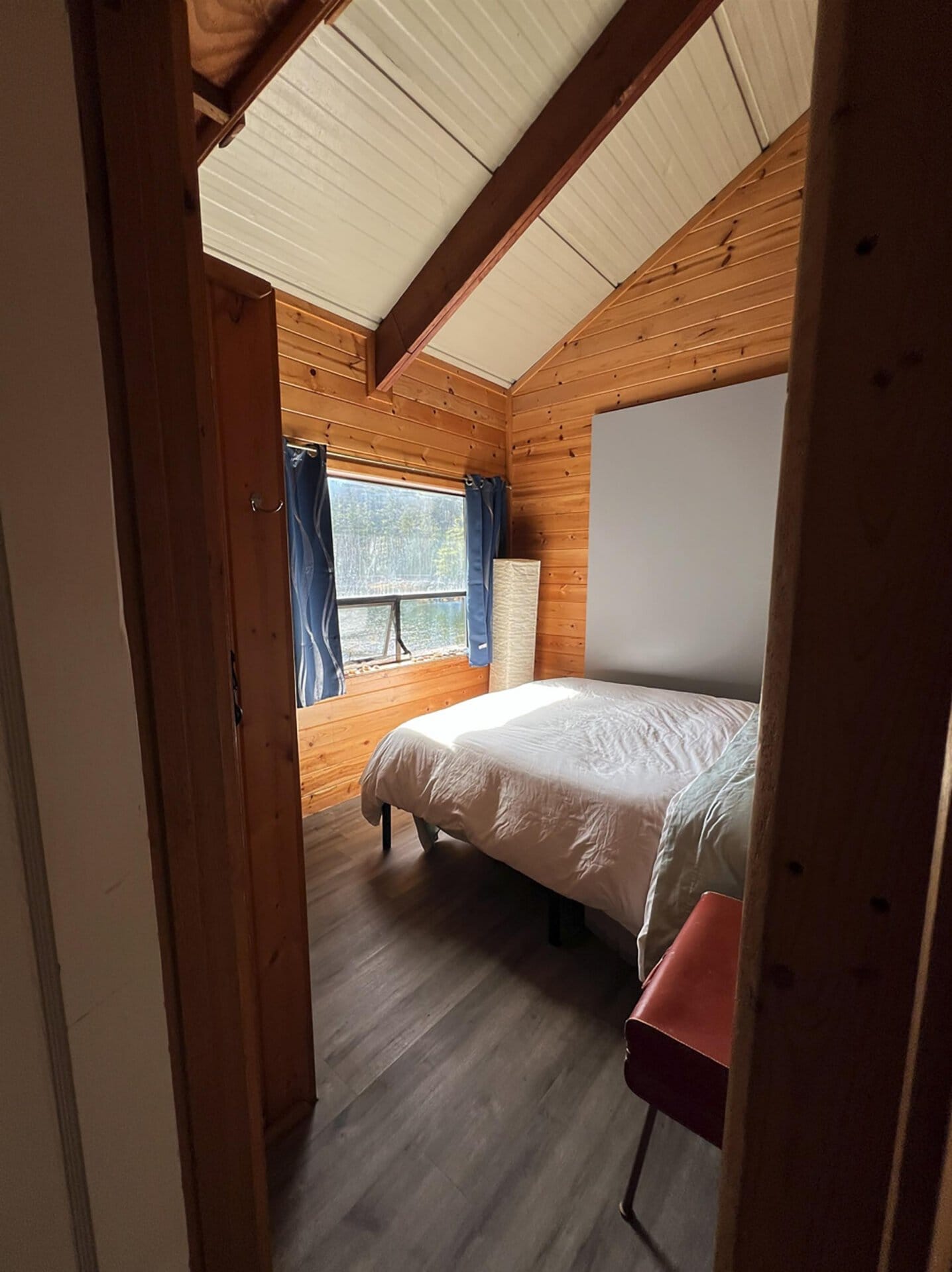 Rustic bedroom with exposed wood walls, a white bed, blue curtains by a window, and a red chair in a small cabin room.