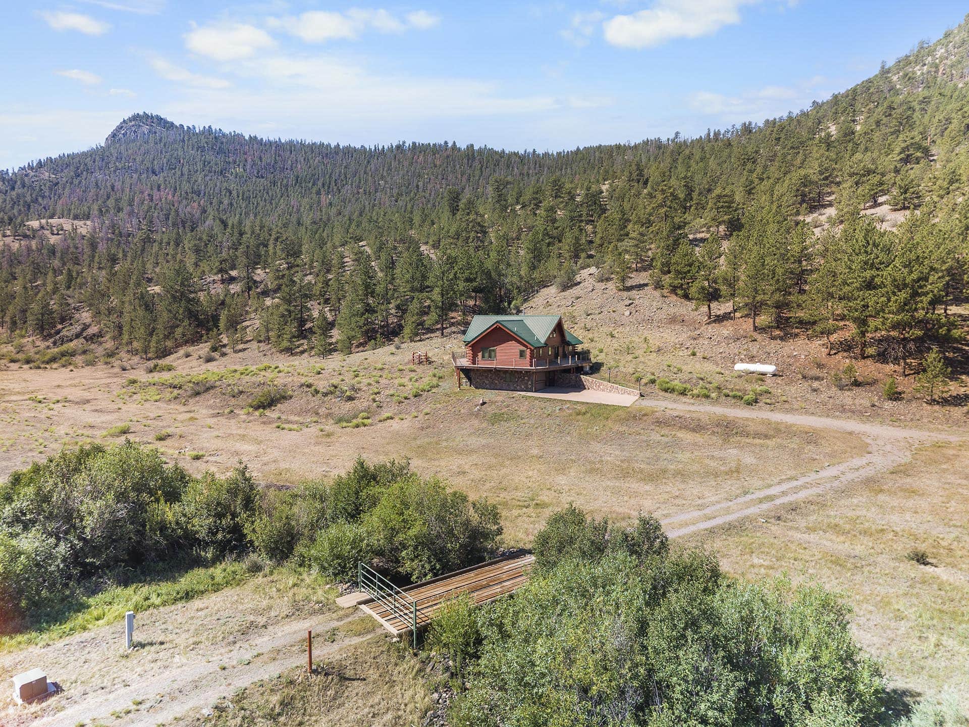 Red wooden cabin with green roof perched on a hillside, surrounded by pine trees and arid grassland; a dirt driveway leads to a raised deck.