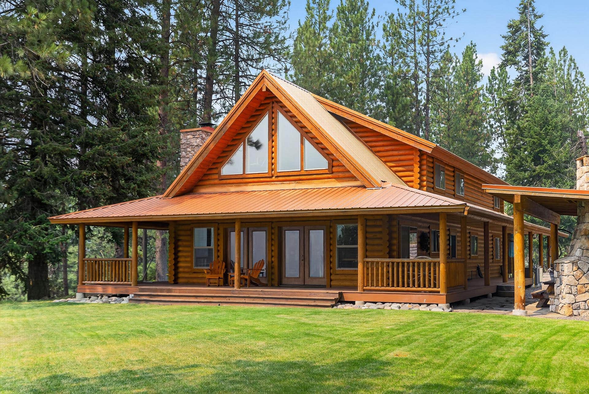 Wood log cabin with a covered porch and triangular attic windows, set among tall pine trees and a green lawn.