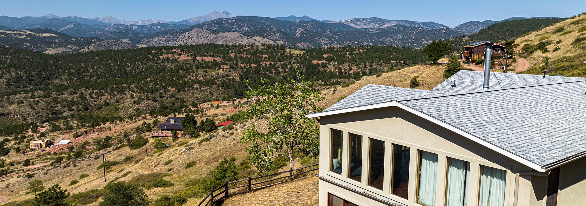 Modern beige house with large windows on a hillside, overlooking a valley and forested mountains.