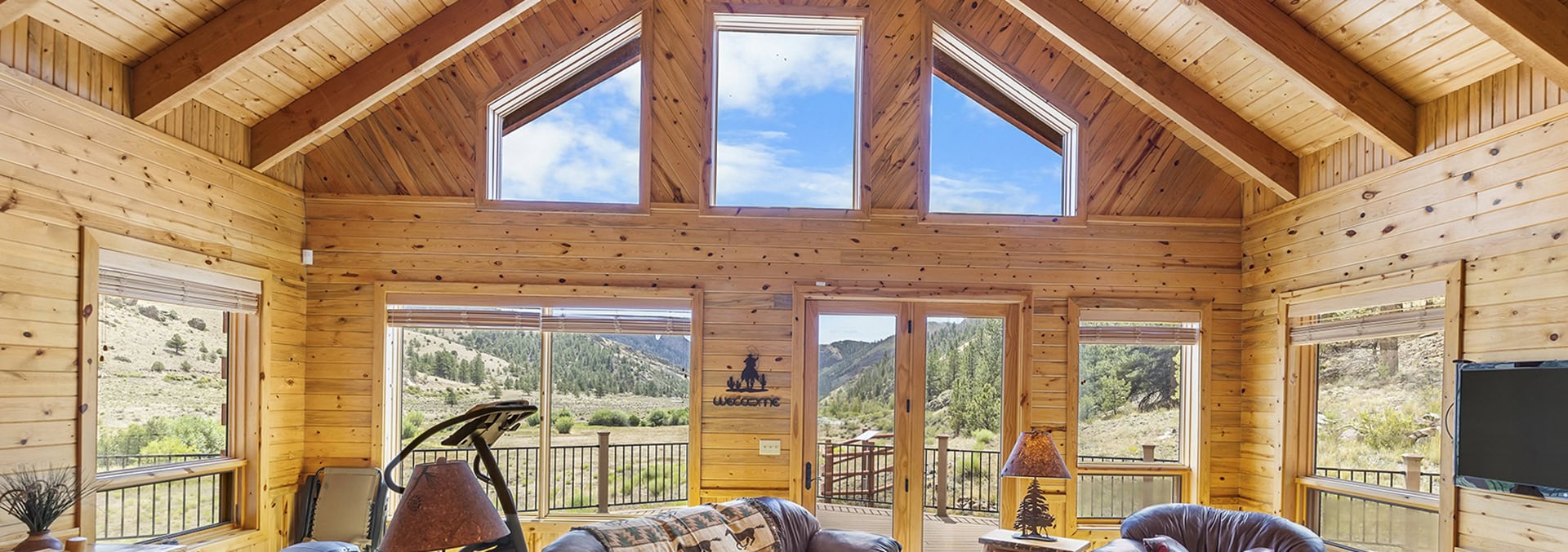 Rustic log cabin living room with a vaulted wooden ceiling and large windows framing a mountain landscape, outdoors visible.