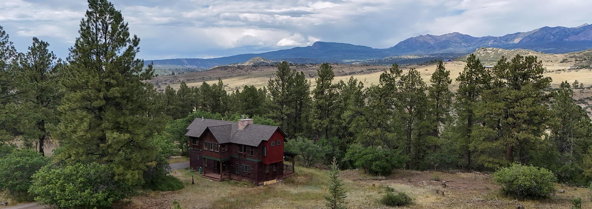 Red two-story wooden cabin surrounded by pine trees in an open meadow with mountains in the distance.
