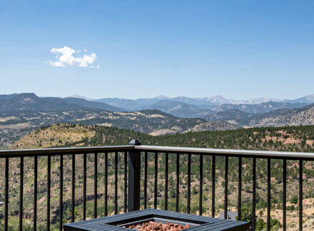 Balcony railing with two wicker chairs facing a mountain valley under a blue sky; a fire pit sits in the foreground.
