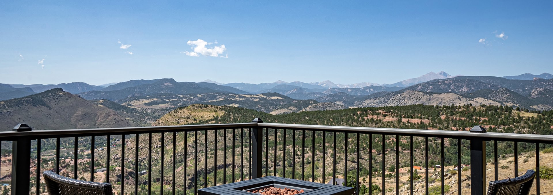 Balcony railing with two wicker chairs facing a mountain valley under a blue sky; a fire pit sits in the foreground.
