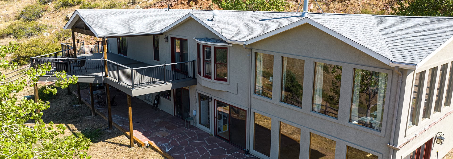 Two-story gray stucco house with a wooden wraparound deck and metal railings overlooking a dry hillside garden.