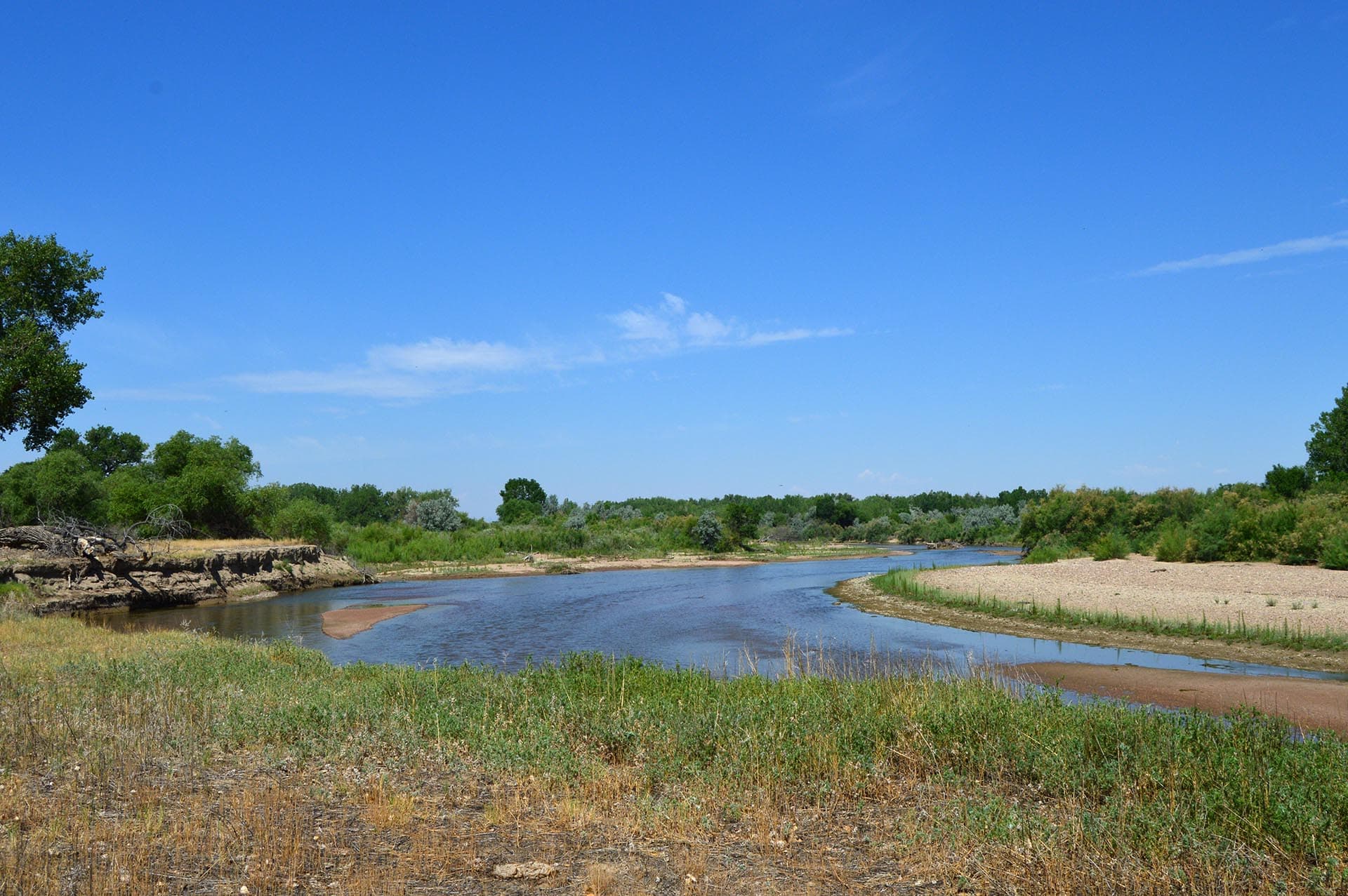 Wide river bend in a dry, grassy riverside under a clear blue sky with green shrubs and trees along the banks