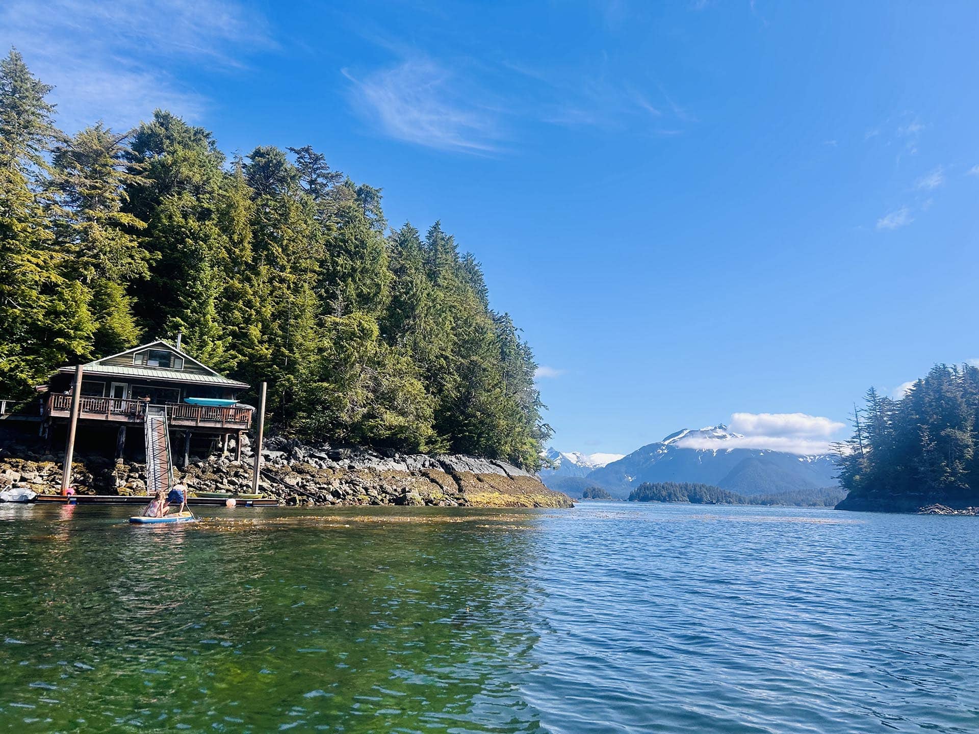 Cabin on stilts along a rocky shoreline, with people paddleboarding in calm green water and dense evergreen trees nearby under a clear blue sky with distant snow-capped mountains.