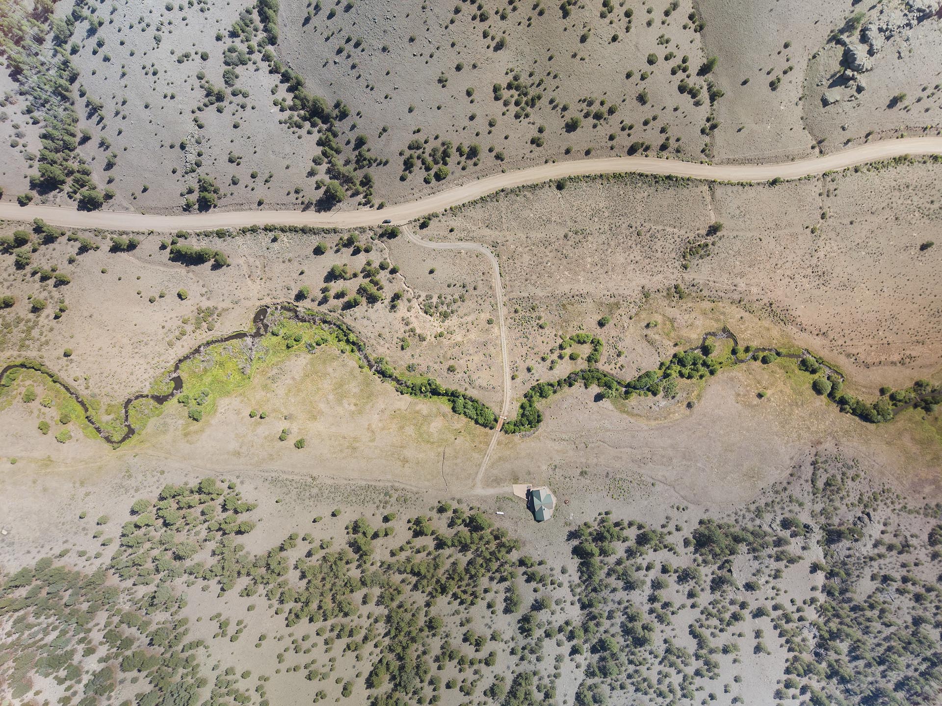 Aerial view of a winding dirt road through a dry landscape with scattered shrubs and a small house near the bottom edge.