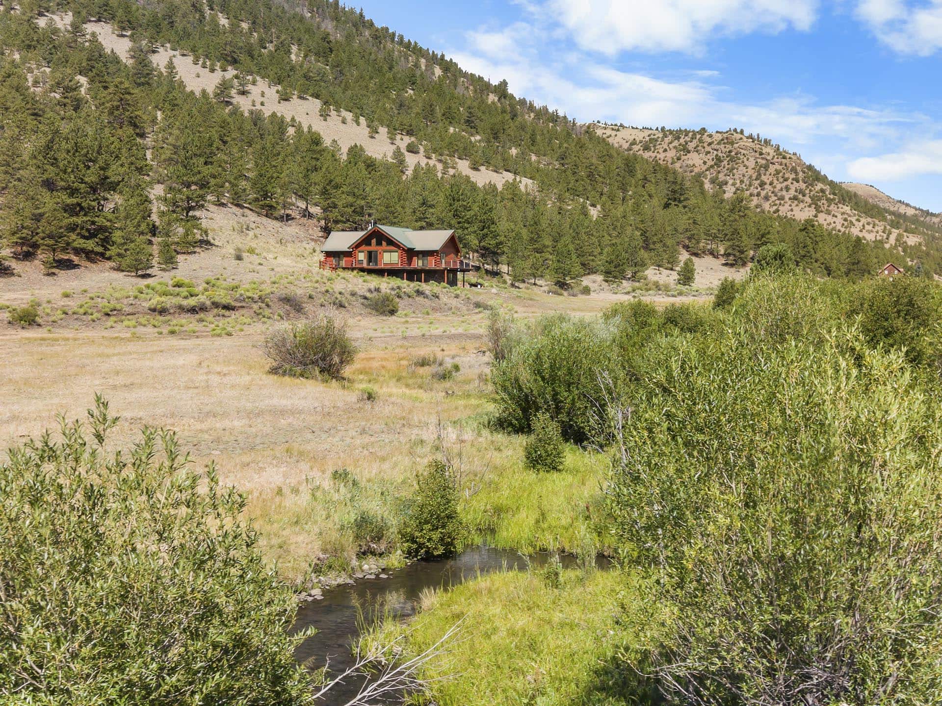 A red cabin on stilts nestled at the edge of a forested hillside with a small stream in the foreground under a blue sky.