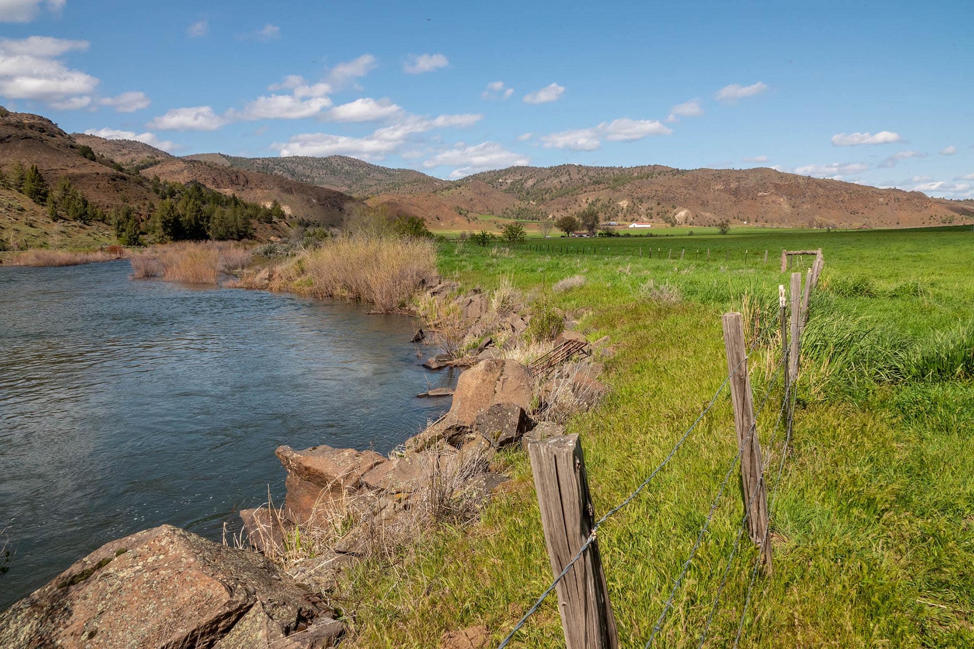 Riverside landscape with rocks, a wooden fence, green fields and distant hills under a blue sky.