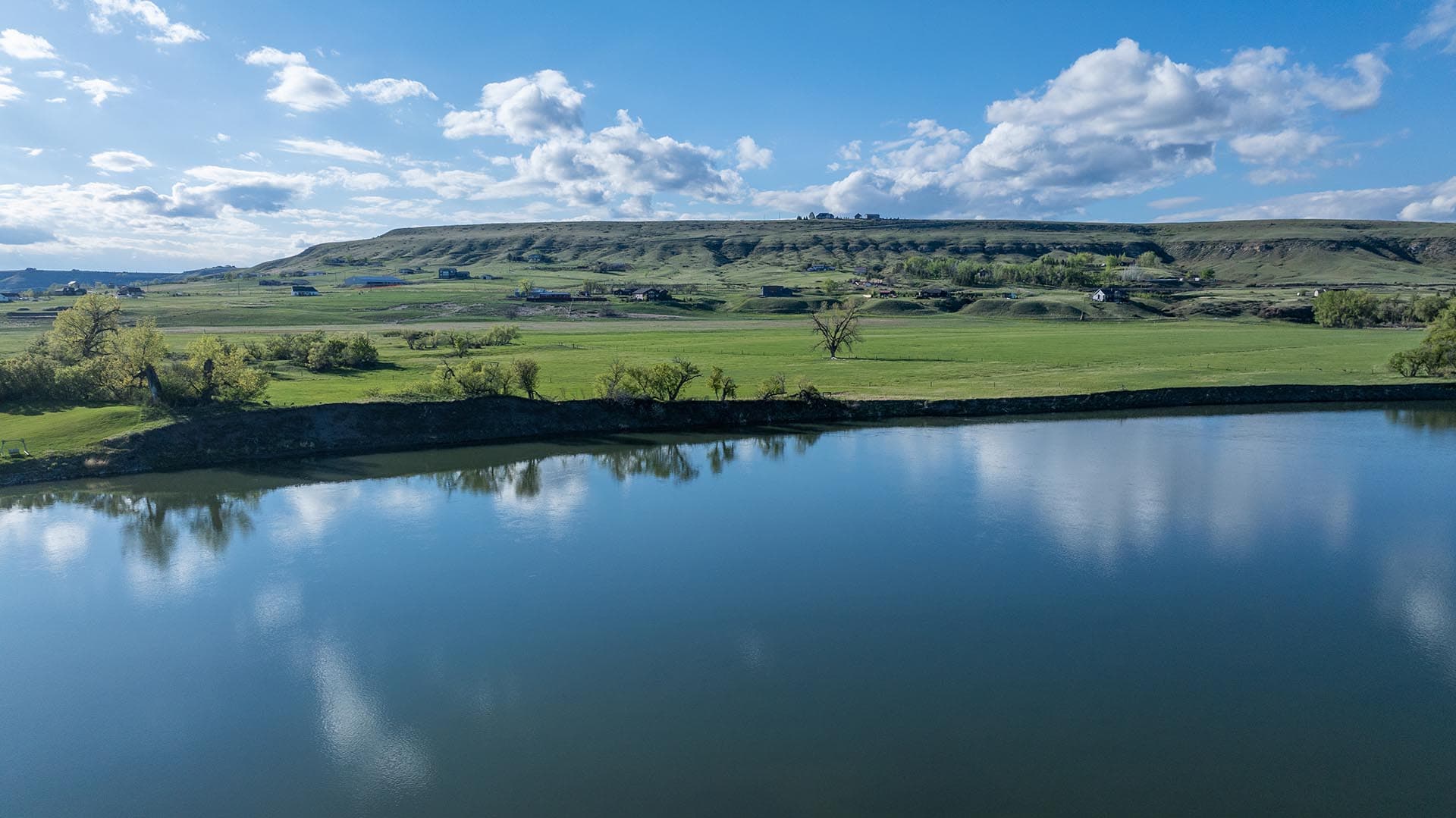 Calm river reflecting the blue sky, with green fields and a low hill dotted with trees in the distance under a cloudy sky.