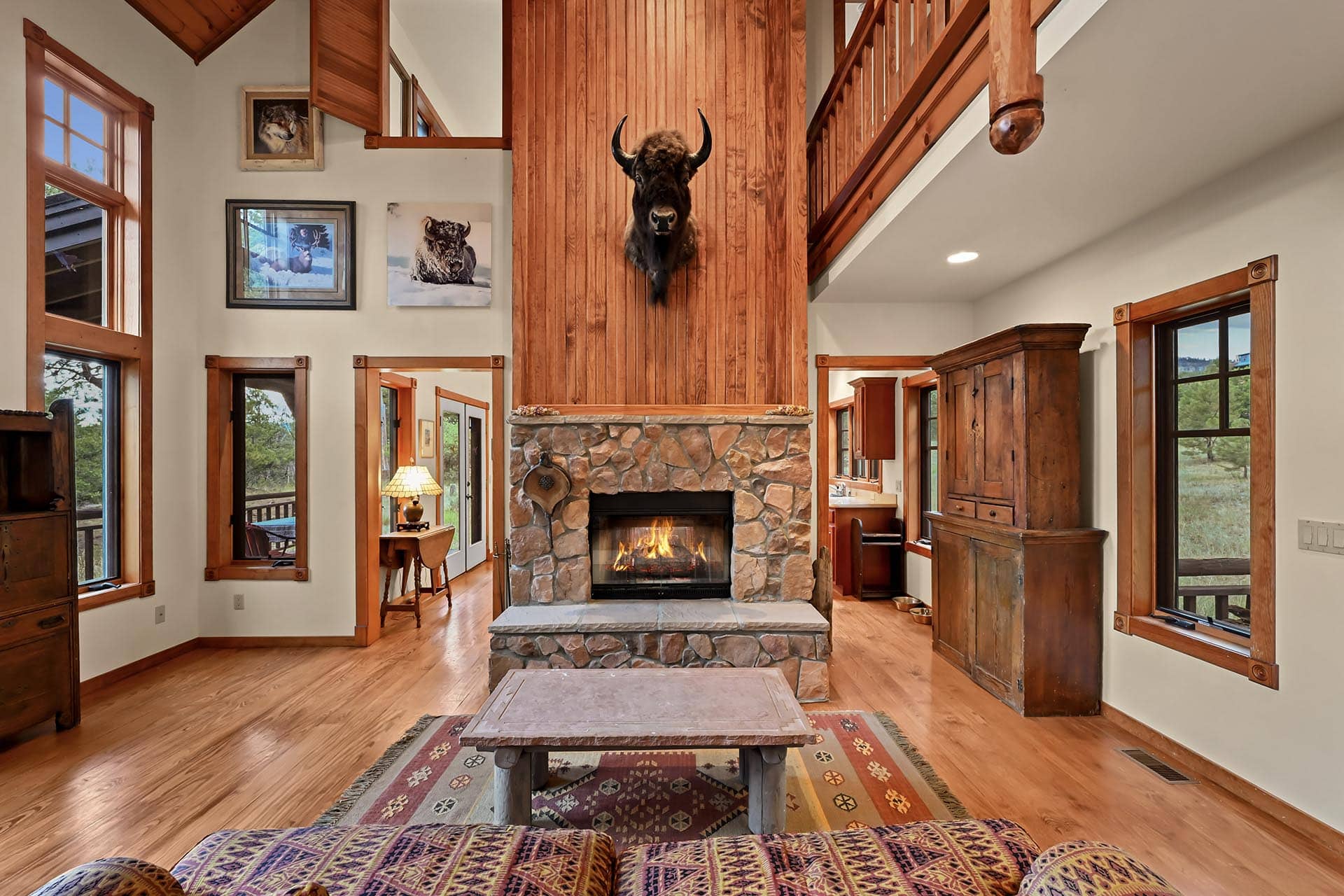Living room with a stone fireplace and a mounted animal head, tall wood paneled wall, and large windows letting in natural light.