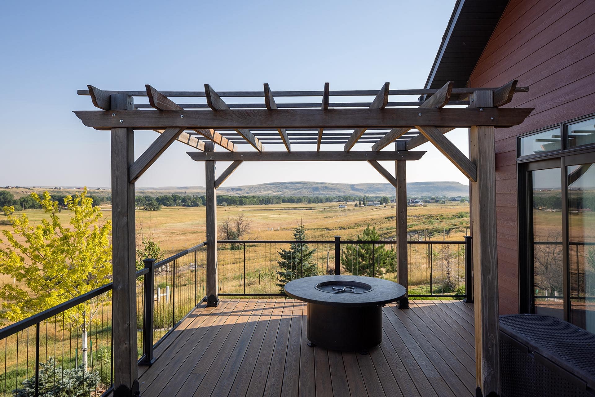 Wooden deck under a rustic pergola overlooking a rural valley and distant hills on a clear day.