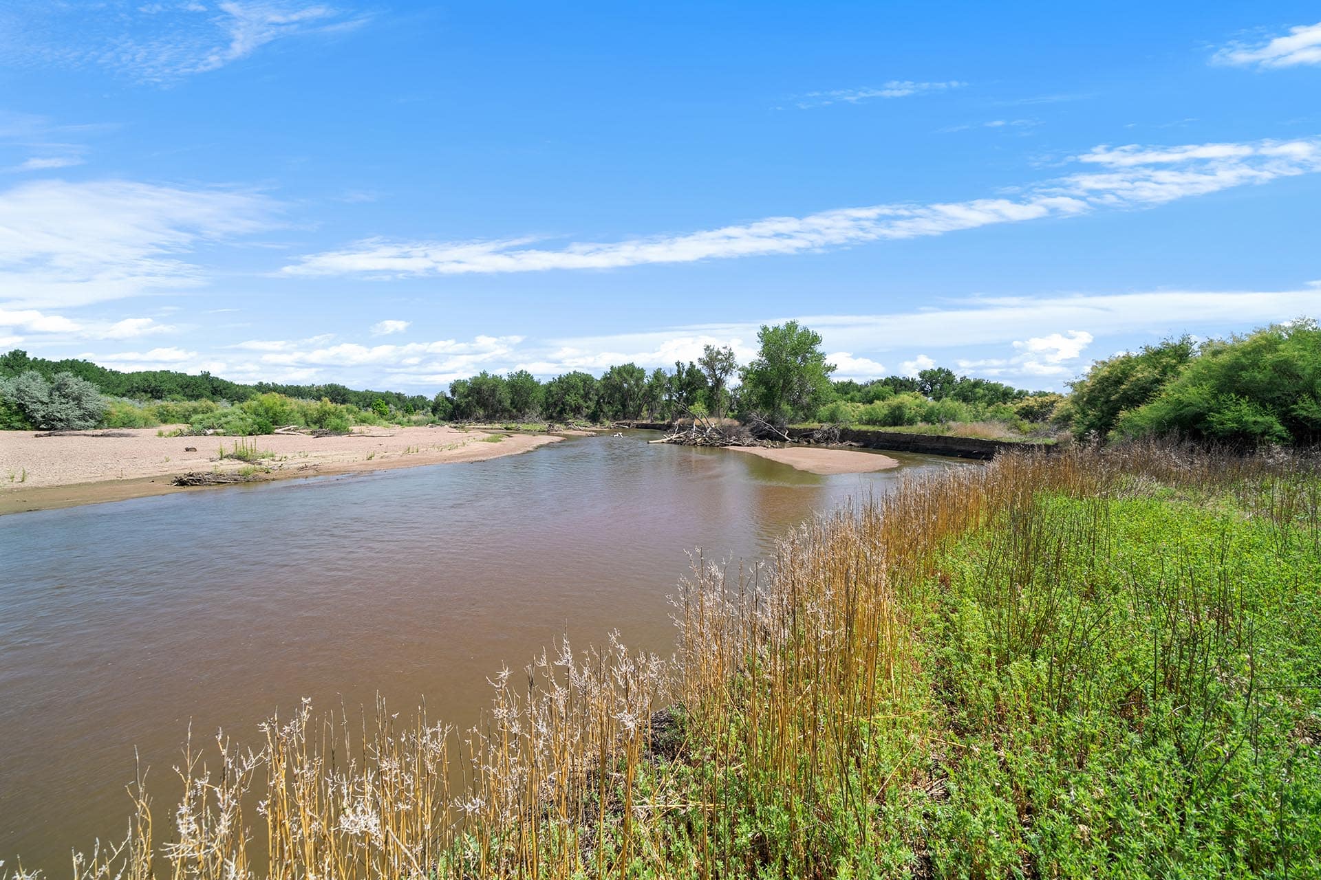 A calm river curves through a sandy banked landscape with green trees and a bright blue sky.