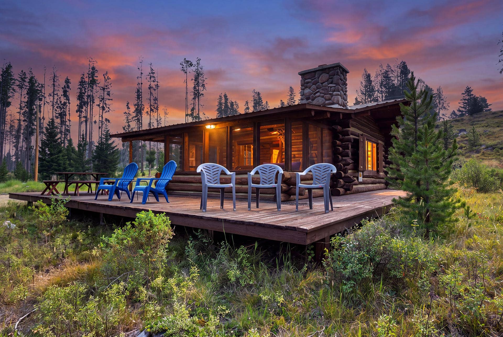 Log cabin with a stone chimney on a wooden deck, orange glow from windows, and colorful plastic chairs facing a sunset forest clearing.