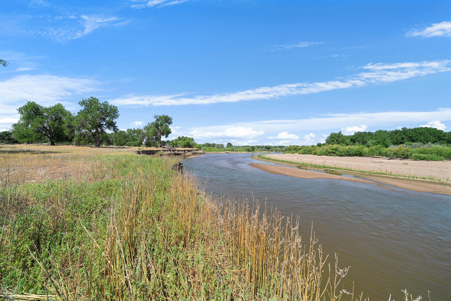 Winding river skirts a grassy riverside under a bright blue sky with scattered clouds and distant trees.