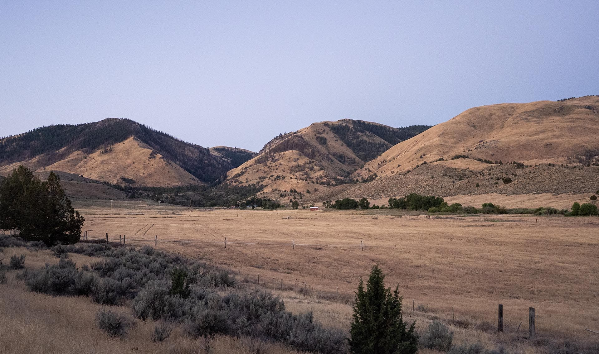 Vast dry valley with brown hills and sparse vegetation, a wooden fence in the foreground, under a clear blue sky at dusk.