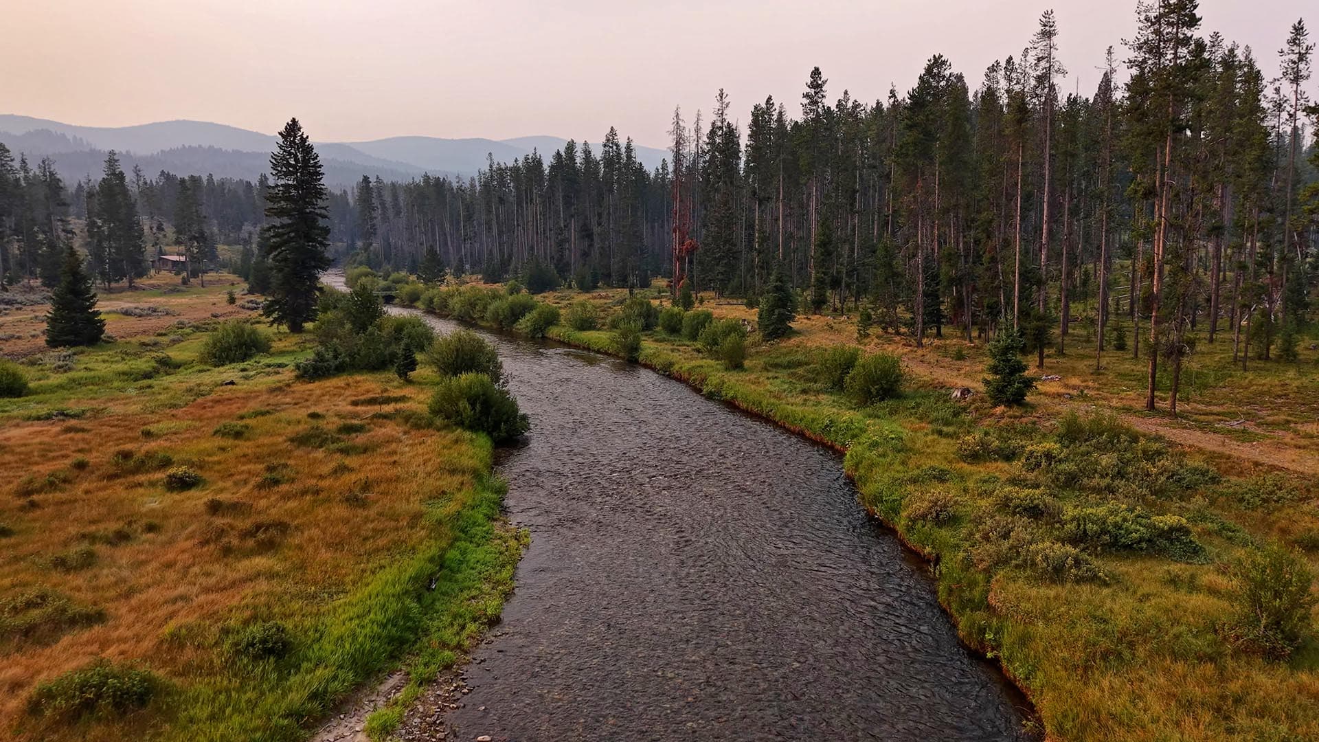 River winds through a grassy meadow bordered by trees with distant hills at sunset/sunrise.