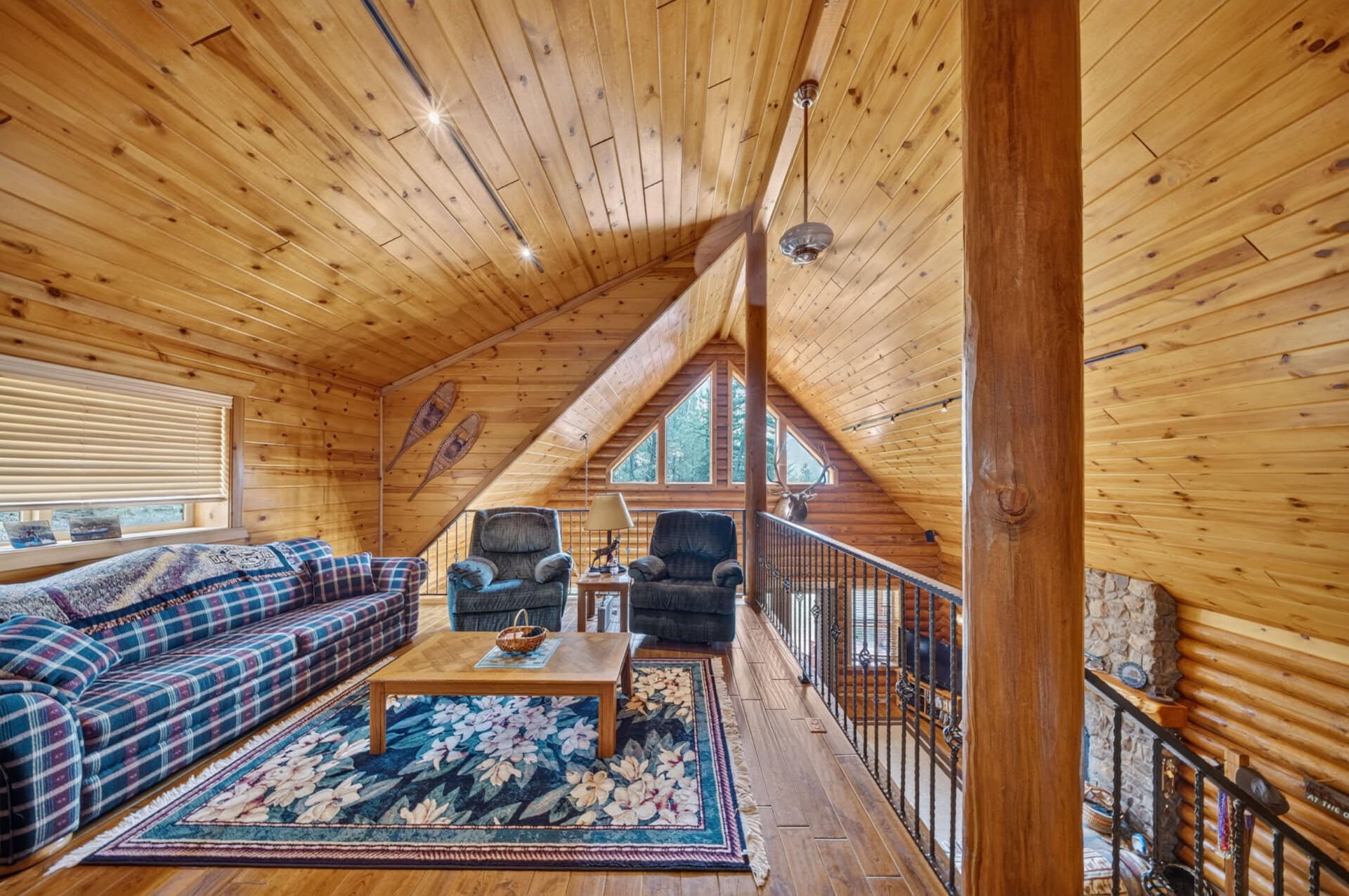 Cozy log-cabin living room with pine paneling, vaulted ceiling, plaid sofa, two armchairs, and a colorful floral rug.
