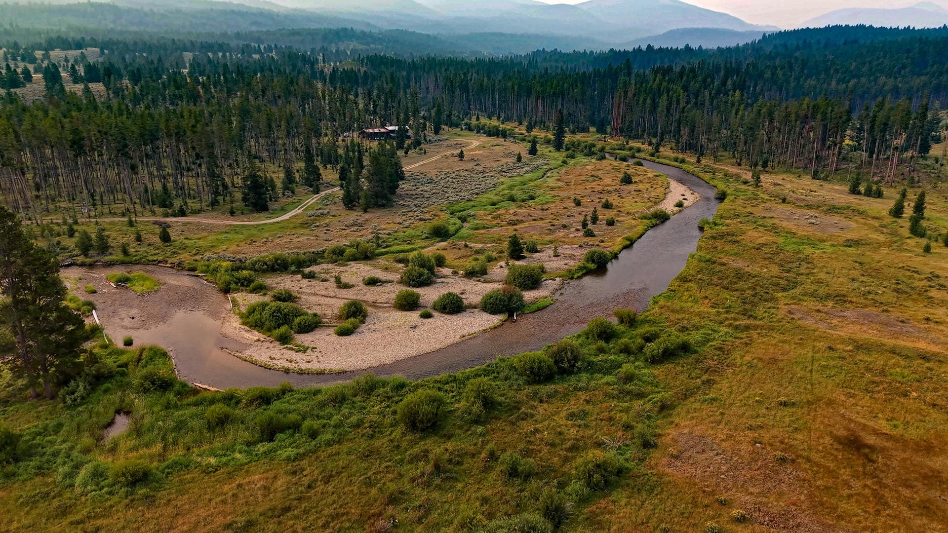 Aerial view of a winding river through a grassy valley with scattered trees and a small cabin in the distance.