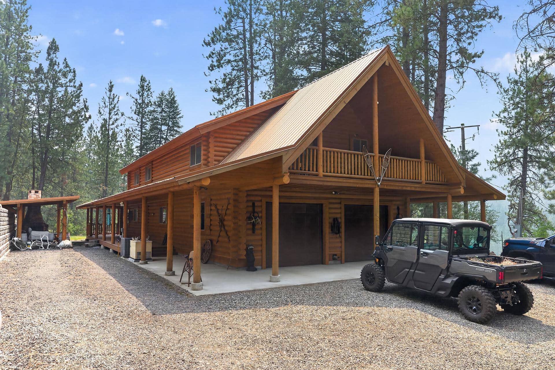 Log cabin with a steep A-frame roof and wraparound porch among tall pines, a gravel driveway, and a dark off-road vehicle in front.