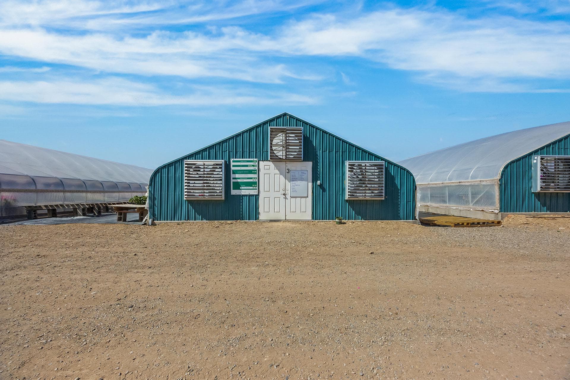 Blue metal greenhouse building with a white door and vented windows in a sunny, arid yard surrounded by other greenhouses under a blue sky.