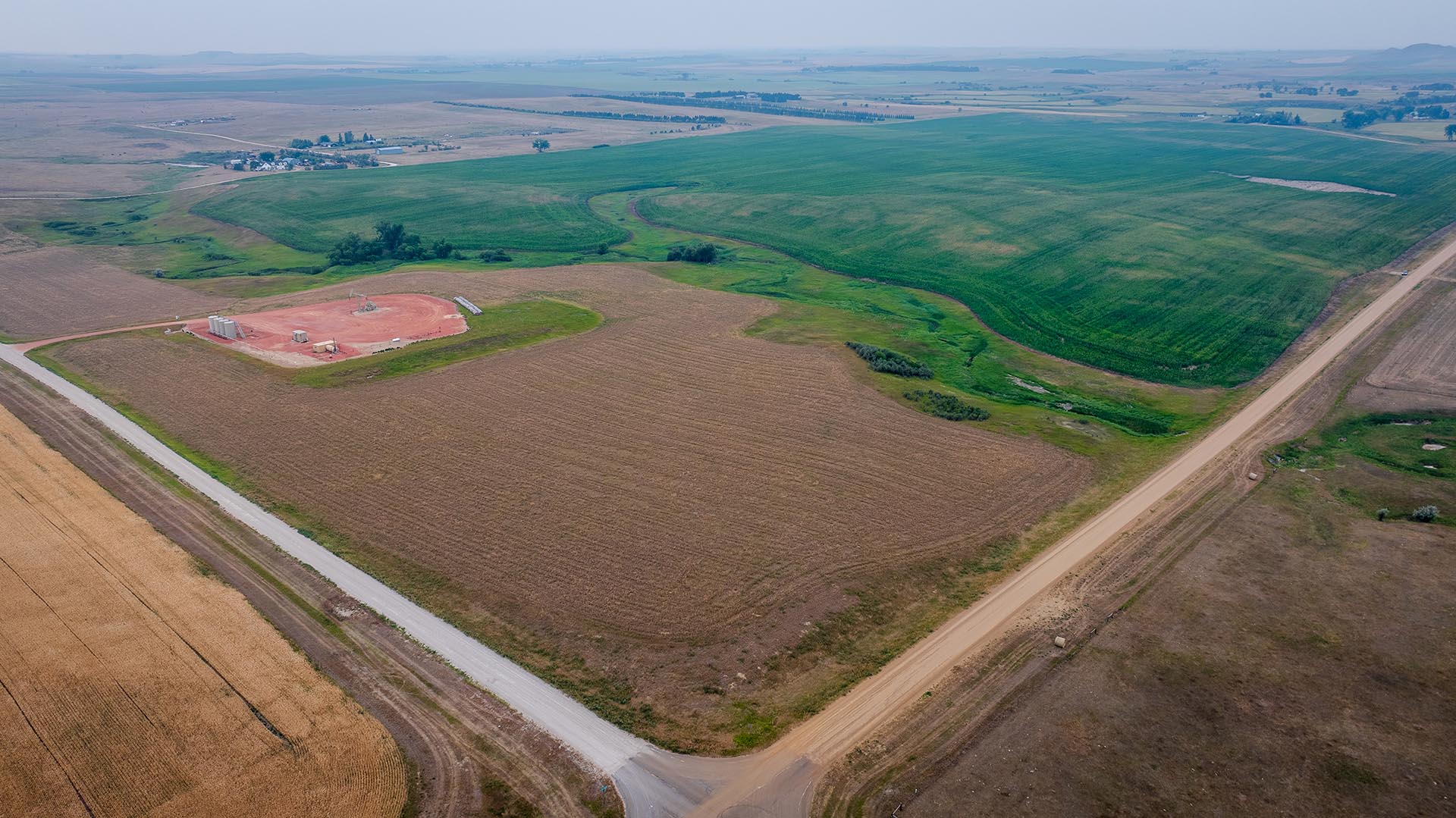 Aerial view of farmland with brown fields, green patches, and a circular red construction pad with equipment near a road edge