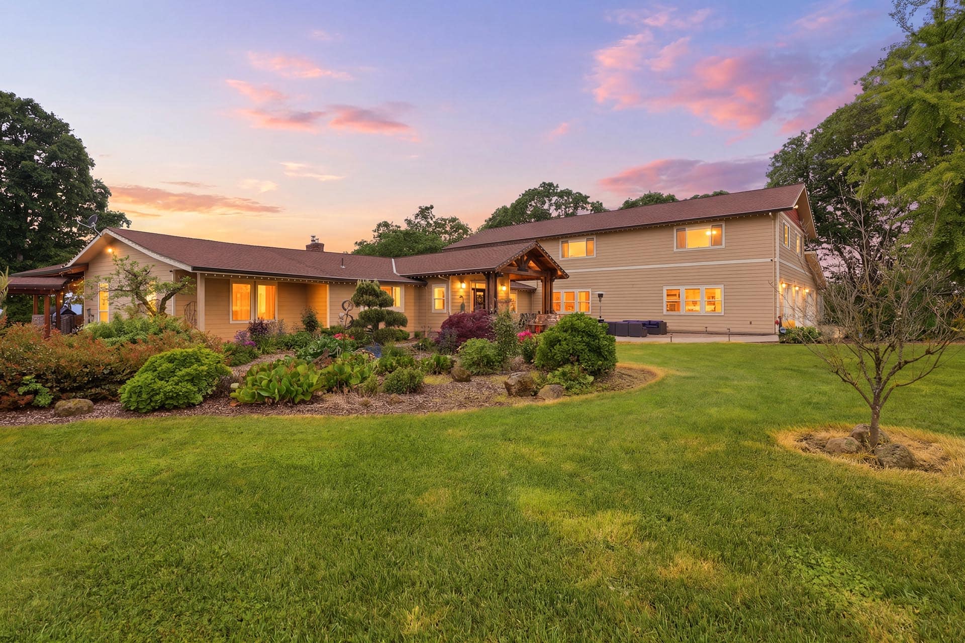 Two-story beige house with a landscaped front yard and warm sunset sky behind it, evening lights glowing in windows