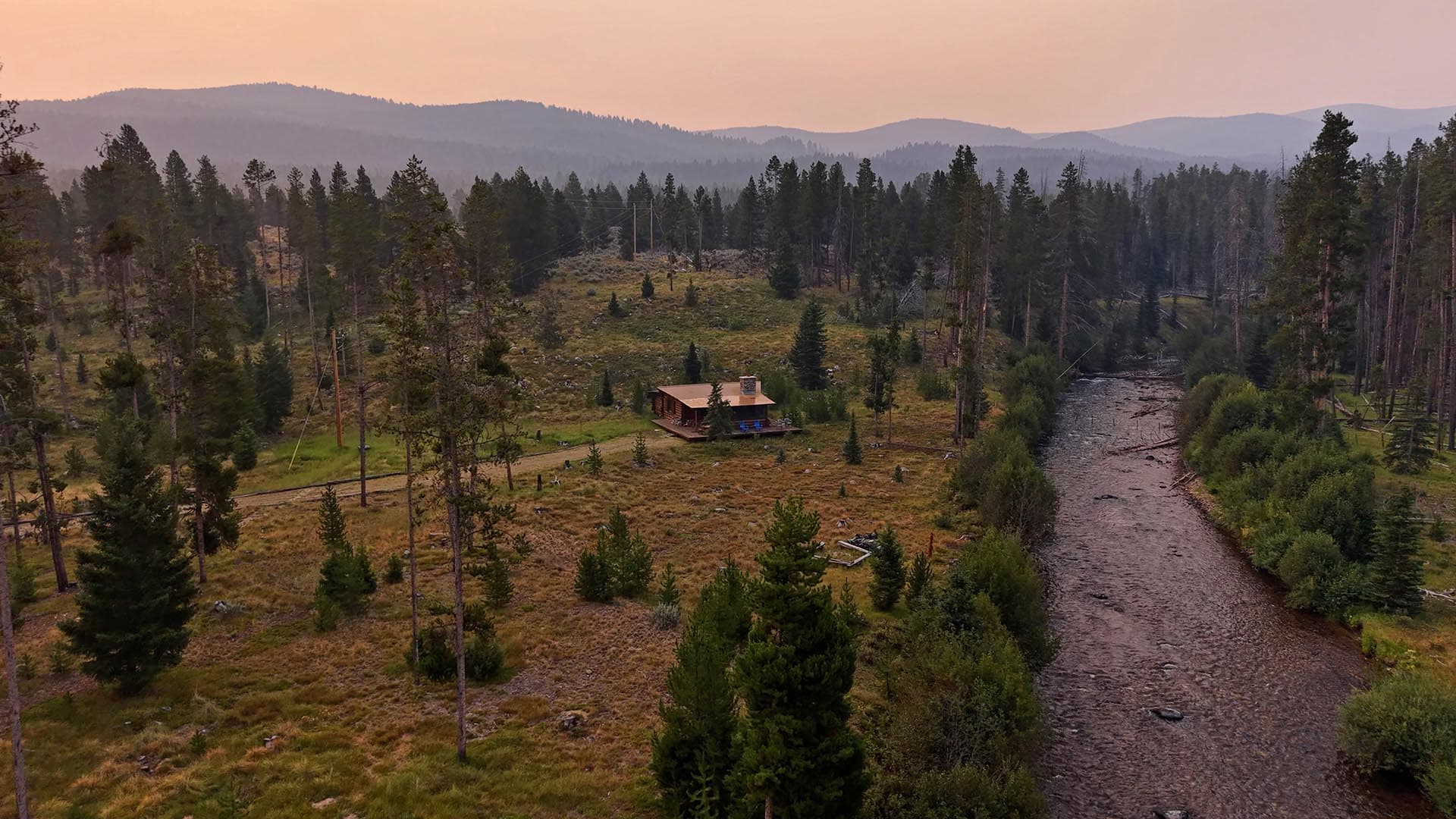 Rustic cabin with a wooden deck sits in a pine forest beside a winding river at sunset.