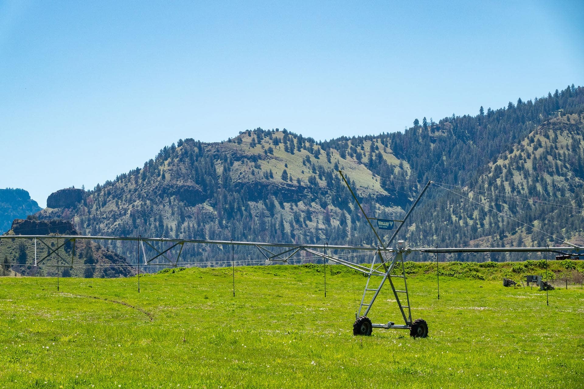 Center-pivot irrigation rig in a green field with forested hills and a blue sky in the background.