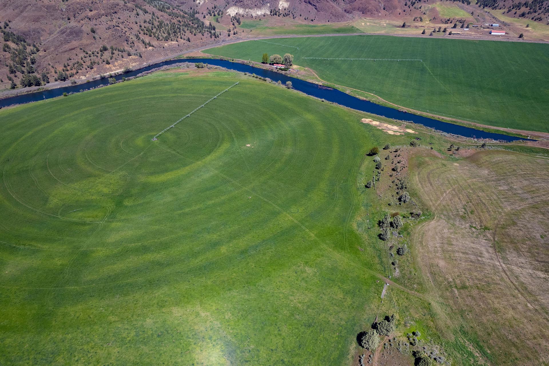 Aerial view of a winding blue river through green farmland with irrigation equipment and distant hills/structures in the background.