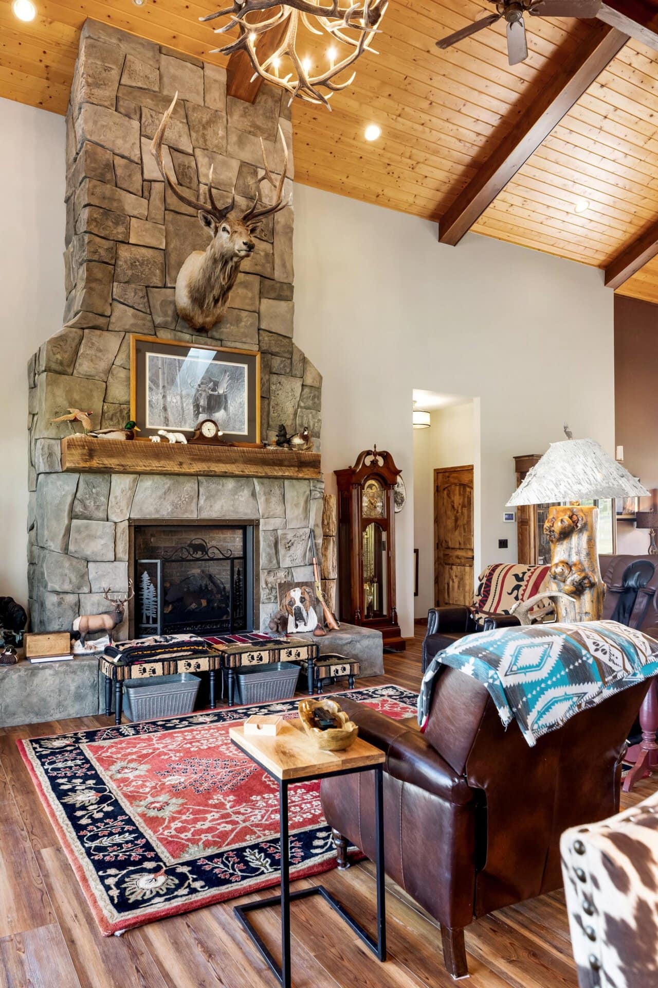 Lodge-style living room with a tall stone fireplace, mounted deer head, antler chandelier, and leather furniture.