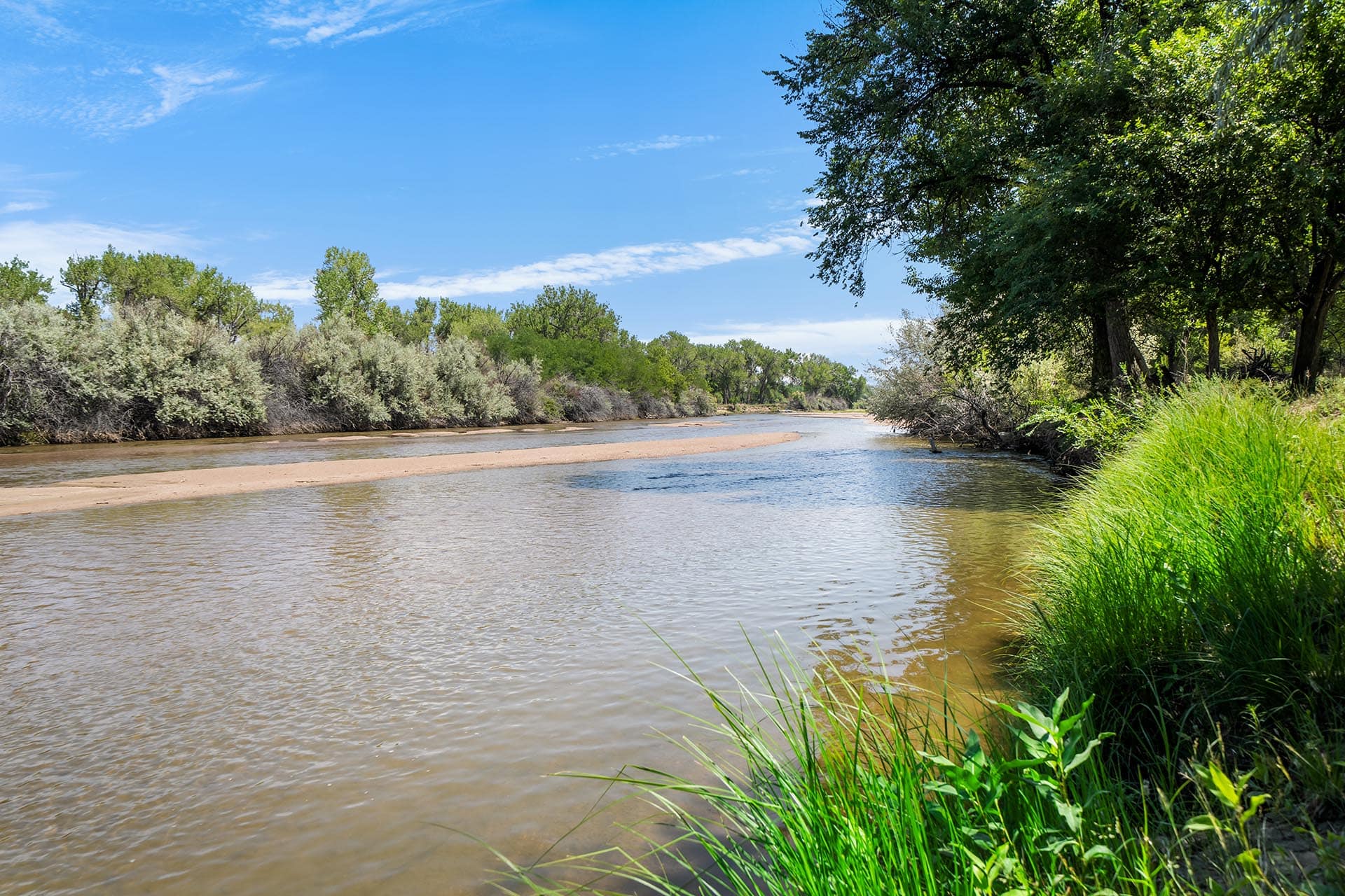 Wide river with sandy banks and green trees along the shore under a clear blue sky.