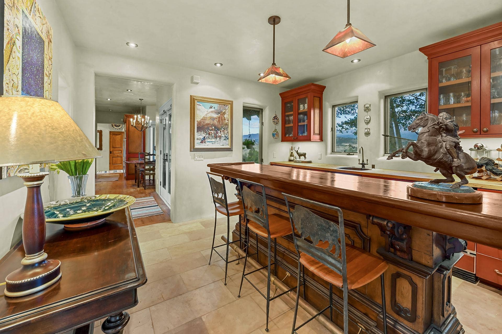 Long wooden kitchen island with bar stools and a bronze horse statue centerpiece in a bright, traditional dining area plus red cabinets and art on the wall