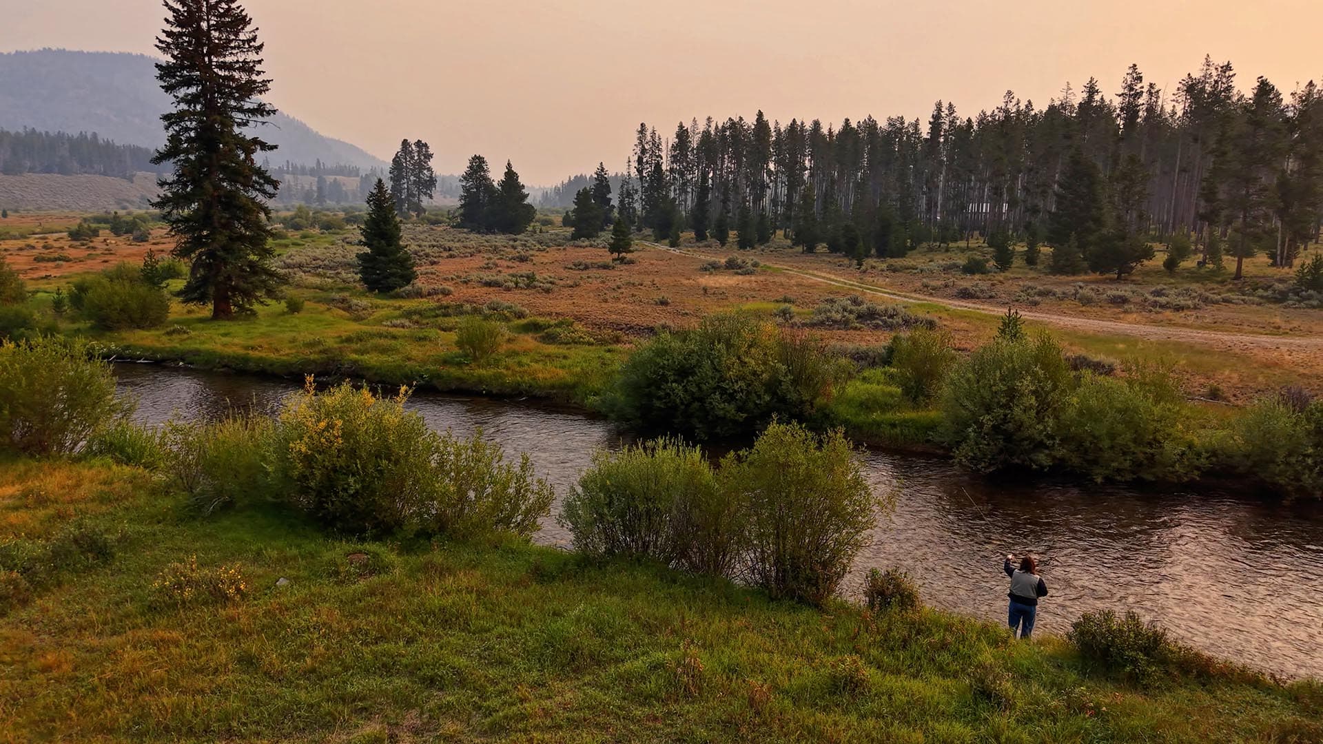 Wide river valley with grassy banks, clusters of trees, and a dirt road under an orange-hued sunset sky; a person stands by the water taking a photo.