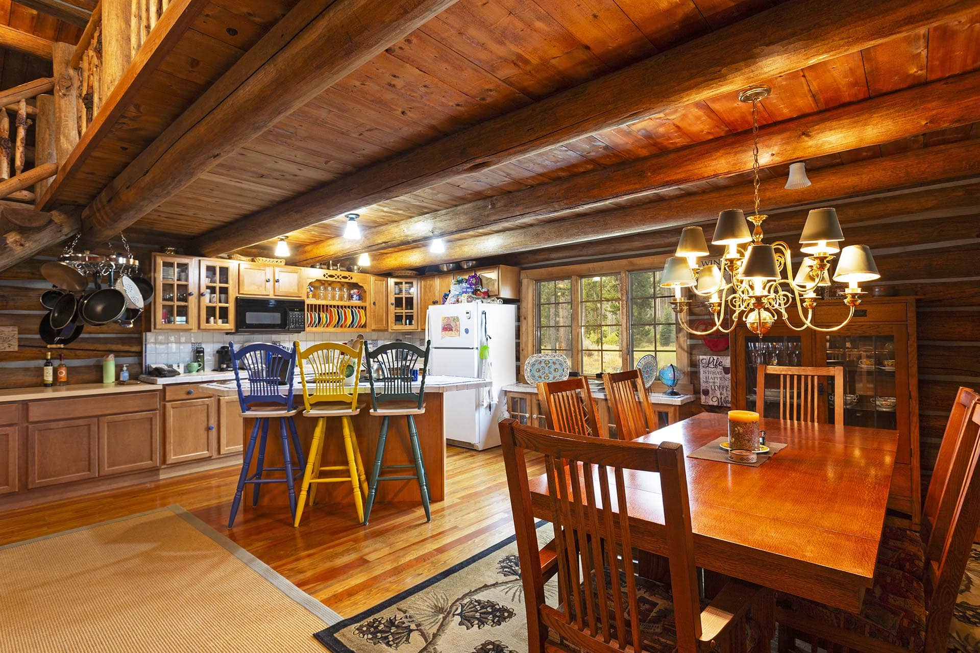 Rustic log cabin kitchen and dining area with exposed wooden beams, island seating on colorful stools, and a brass chandelier.
