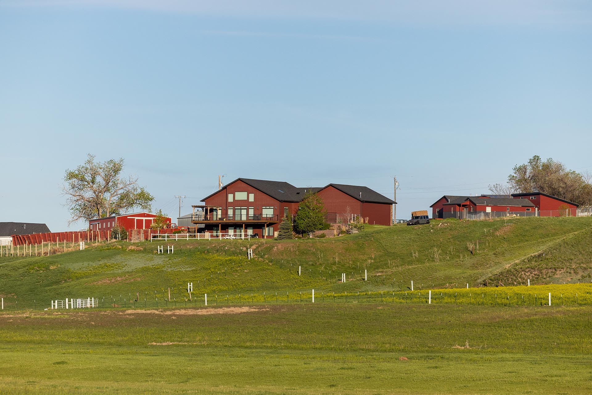 Red farm buildings on a gentle hill with white fences, a decked house, and trees against a clear blue sky.