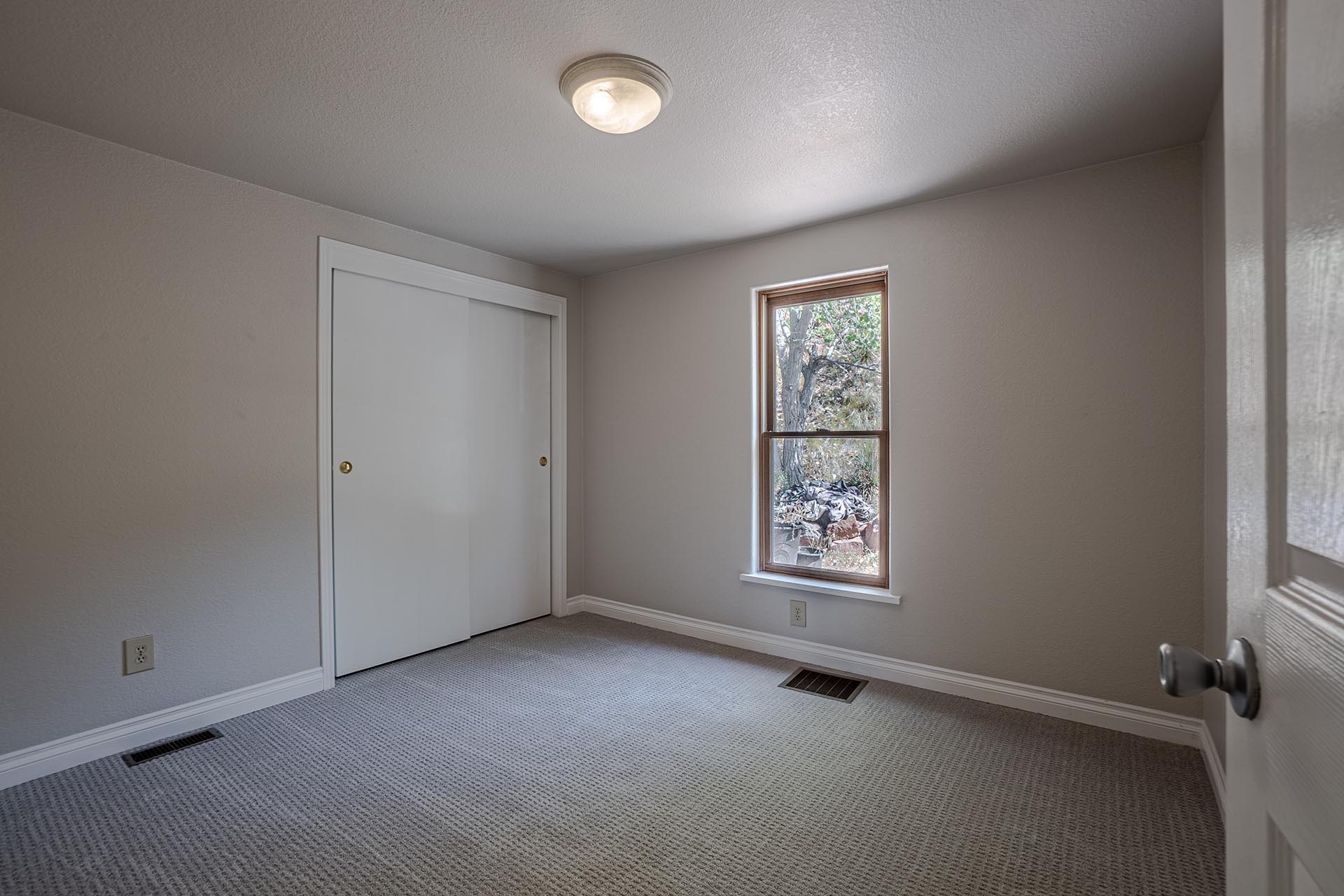 Empty beige bedroom with white sliding closet doors and a small window to the right; carpeted floor and a ceiling light.