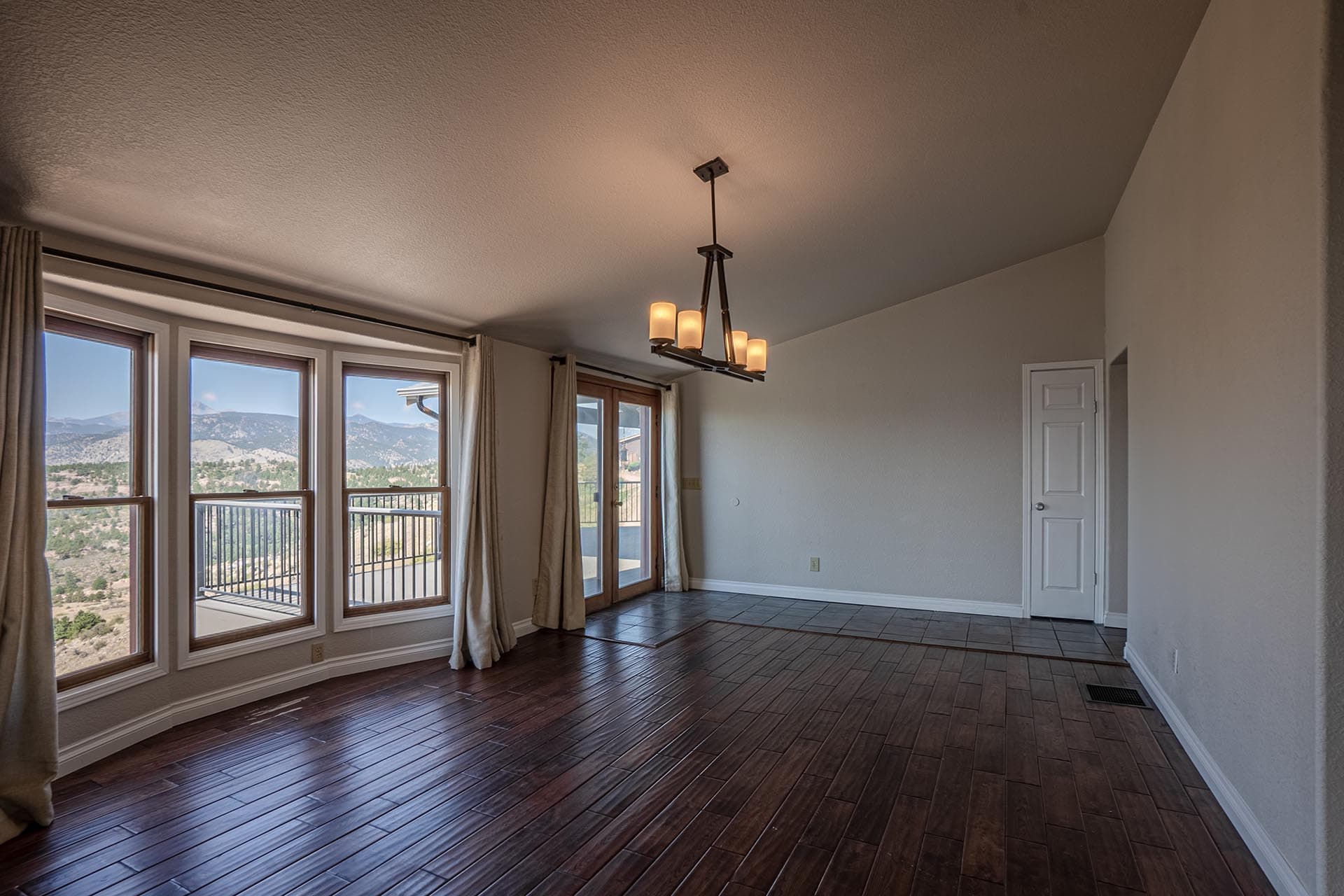 Living room with dark wood flooring, large windows showing a mountain landscape and a balcony railing, plus a modern chandelier hanging from the ceiling.